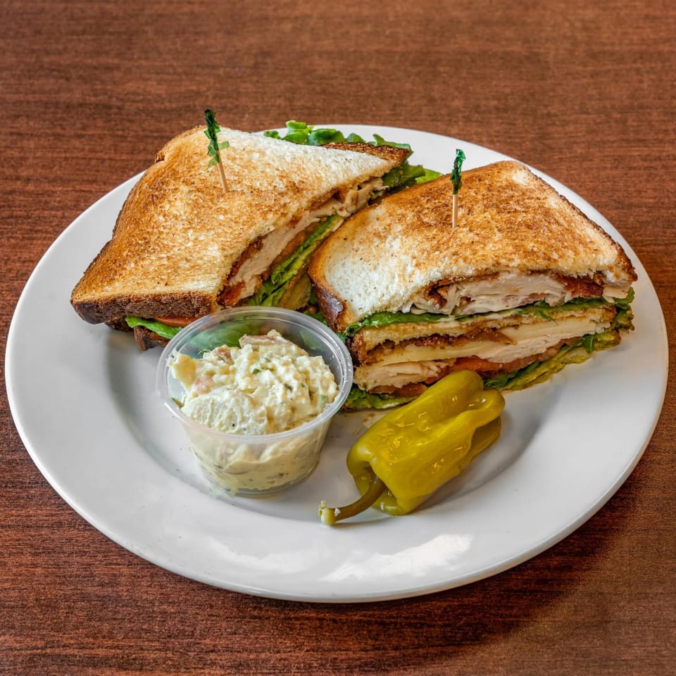 Club sandwich with toasted bread, lettuce, tomato, and meat, served with potato salad and a pepper.