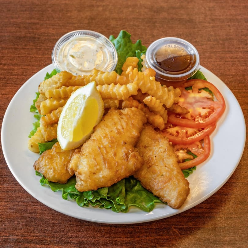 Crispy fish and chips with a lemon wedge, lettuce, tomato slices, tartar sauce, and ketchup.