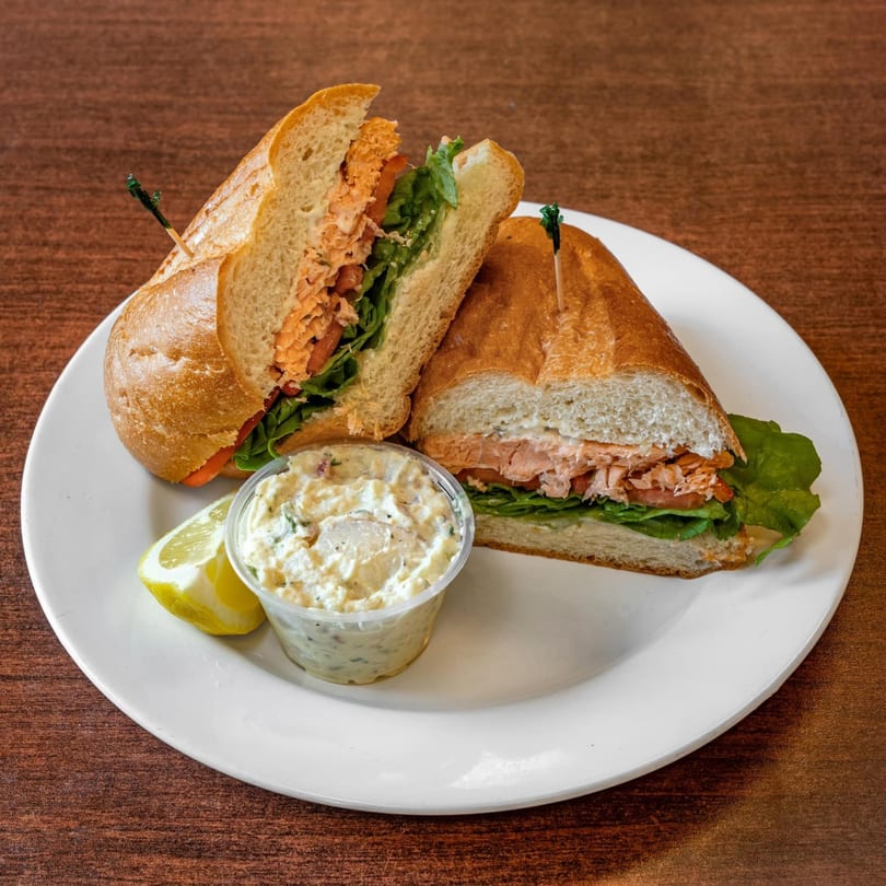 A salmon sandwich on a baguette with lettuce and tomato, served with potato salad and a lemon wedge.