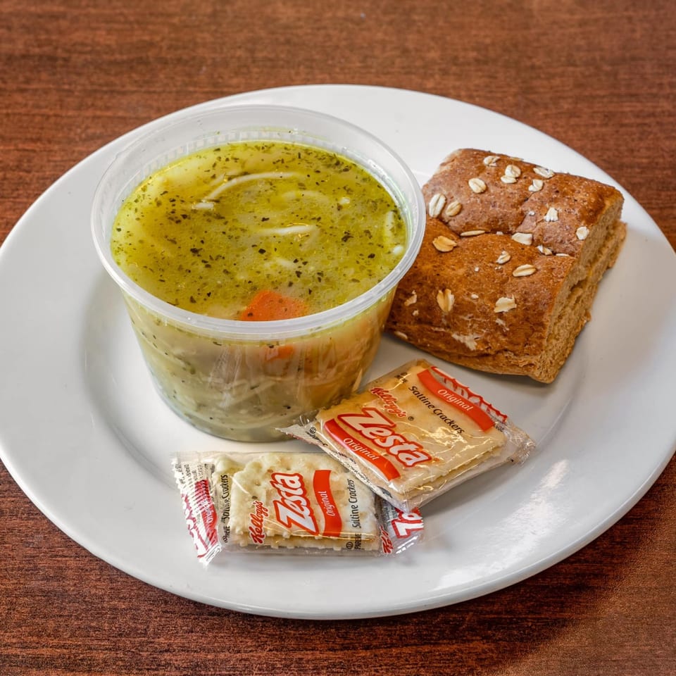 Chicken noodle soup in a plastic container, with a side of whole wheat bread and Zesta crackers.